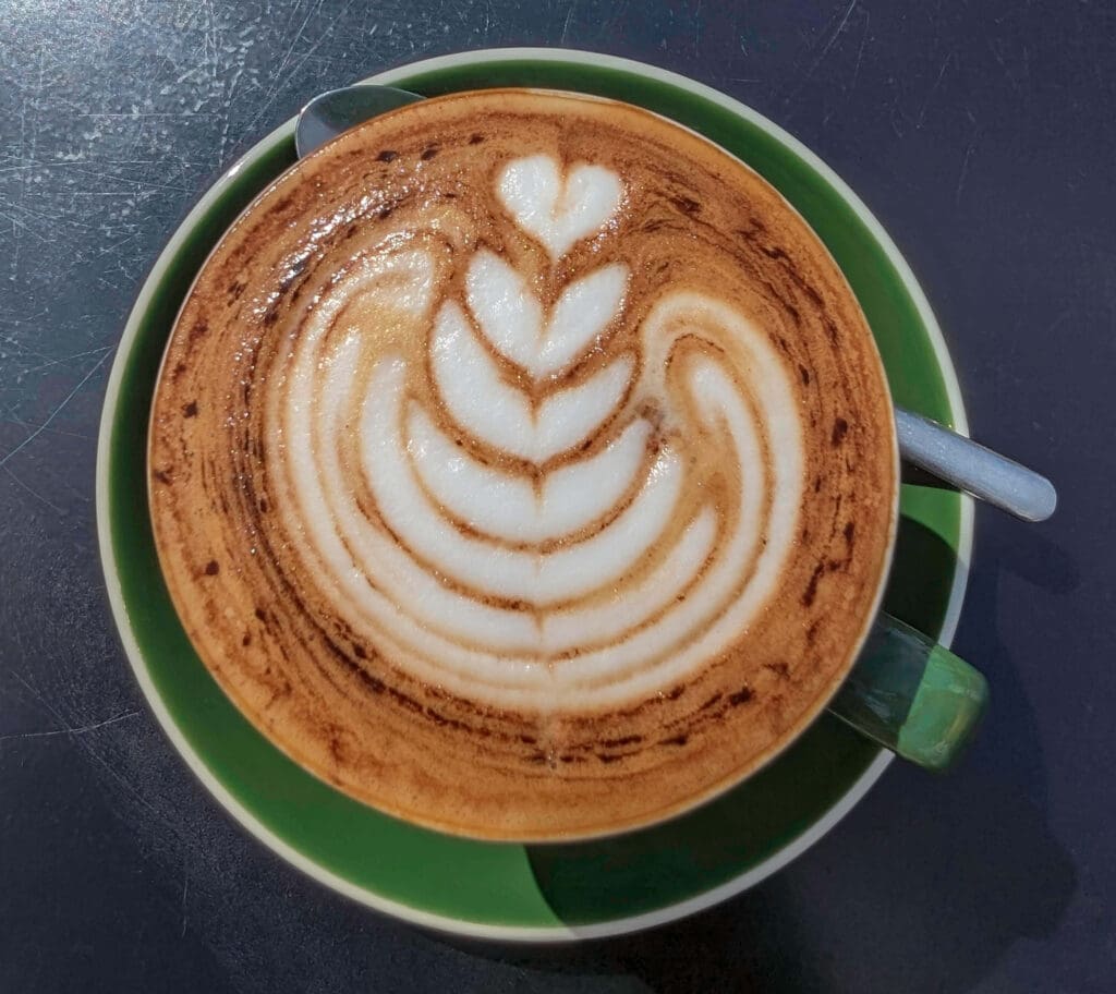 A caramel-colored latte with a beautiful white foam flower design on top all inside a green coffee cup