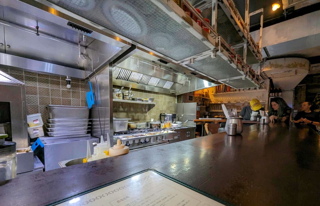 A silver clean kitchen with three people sitting at the bar area of the Berenjak restaurant in London