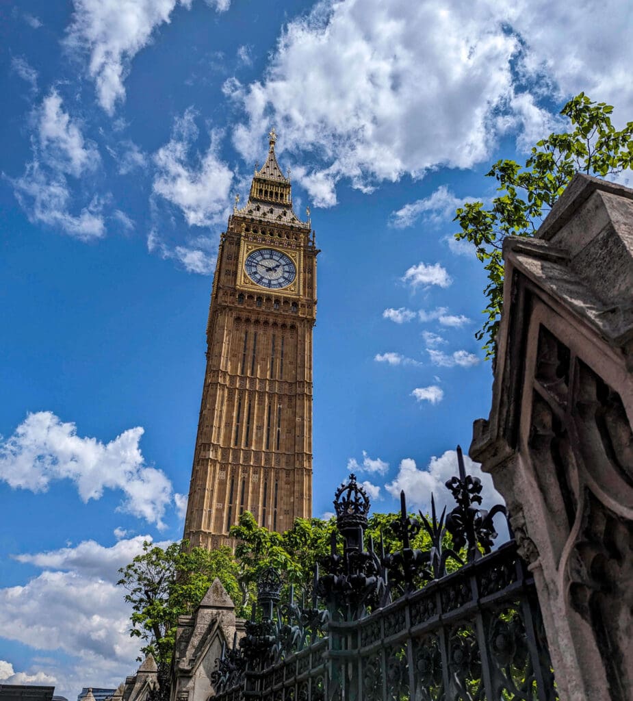 Looking up at the big ben clock tower reaching towards the sunny blue sky with trees and an old gate in the foreground