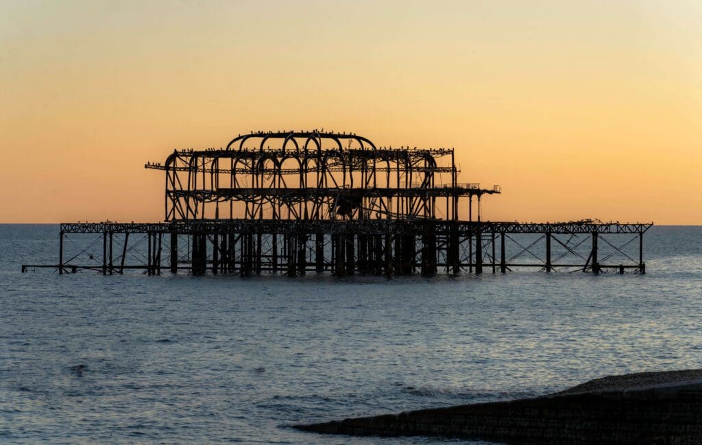 A warm orange and yellow sunset backgrounds the ocean and burned remains of the old West Pier