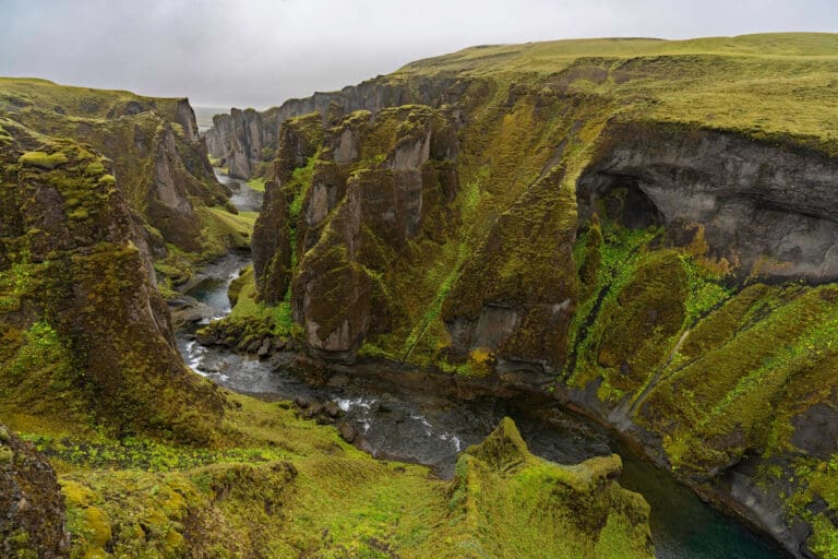 Dark water calmly flows through the otherworldly Fjaðrárgljúfur gorge coated in multi-colored moss