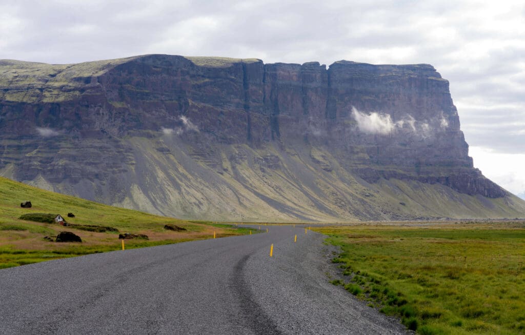 A black dirt road winding through Icelandic grasslands with a half-buried house and giant plateau in the background