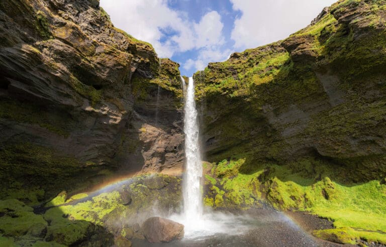 Water flows over the green mossy overhang of the Kvernufoss waterfall as a small rainbow shines across the bottom