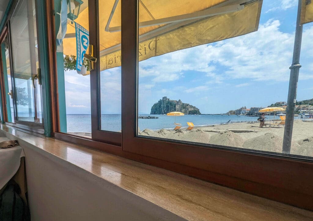 Ocean waters washing onto a sandy beach with the Castello Aragonese d'Ischia in the distance looking from Ol Sole Mio