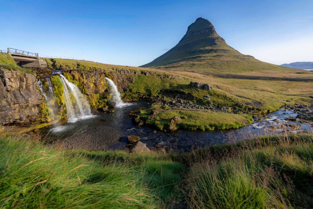 Soft flowing waters of the Kirkjufellsfoss waterfall featuring long wispy grass and a dorsal fin shaped mountain