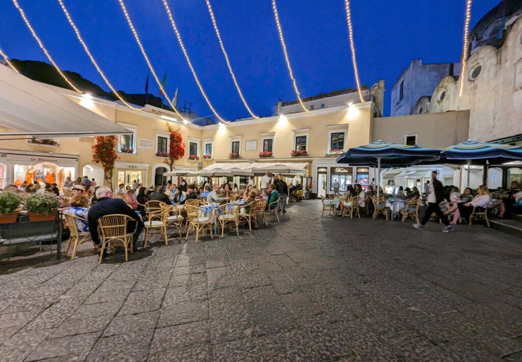 String lights hang overtop people sitting in front of cafes in the Capri Town Square