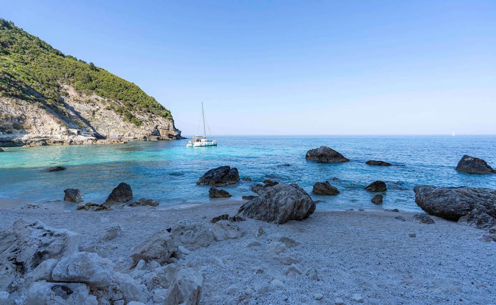 White rocks sit in a shaded beachfront with a boat floating in the quiet cove.