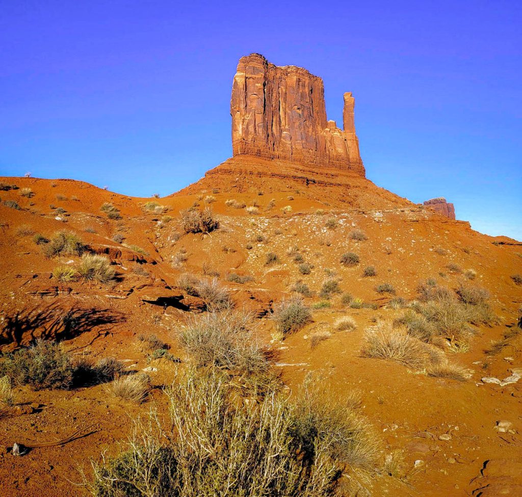 A blue sky and dry gravel surround one of the many stunning buttes in Monument Valley from the Wildcat Trail, one of the best hikes in Arizona.