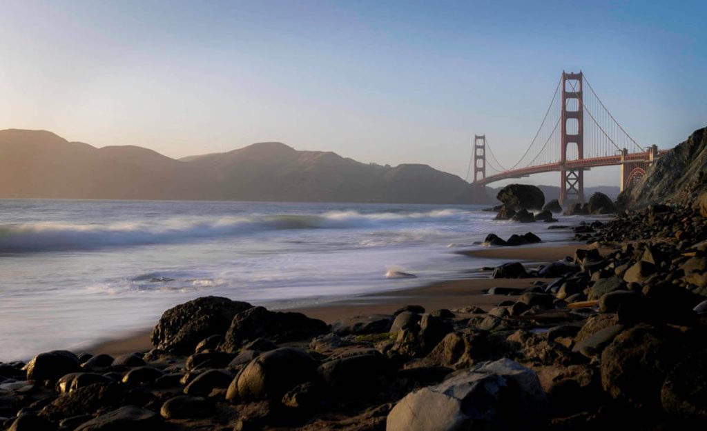 Waves lightly roll onto the Golden Gate Beach as the setting suns paints a golden light onto the distant Golden Gate Bridge.
