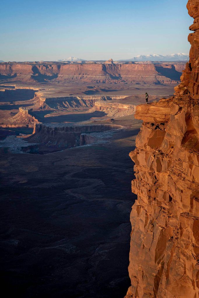 As the sun rises, a girl precariously stands on the edge of the Green River Overlook showing why this is one of the best Canyonlands hikes.