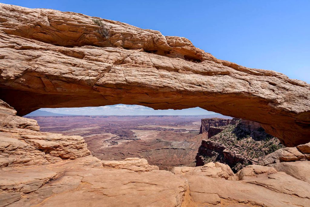 A long curved rock bridge overlooks the expansive Canyonlands vista, as the sun shines overhead.