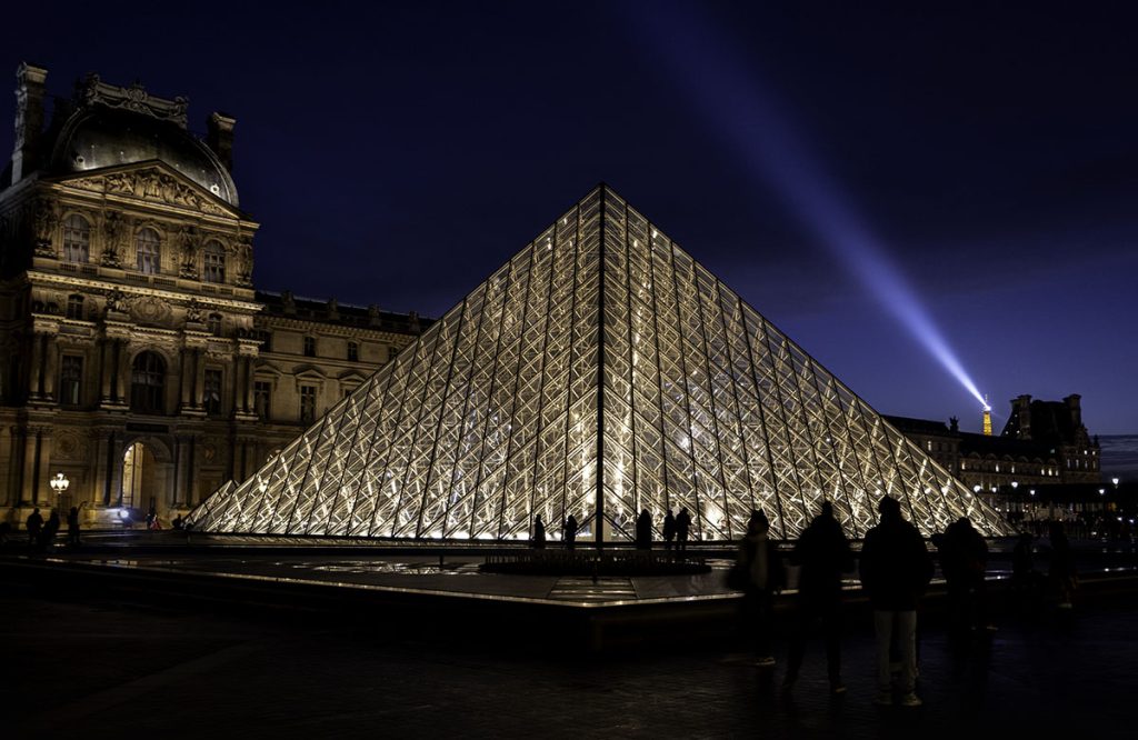 Illuminated pyramid and courtyard shine in the night as the Eiffel Tower view glistens beyond.